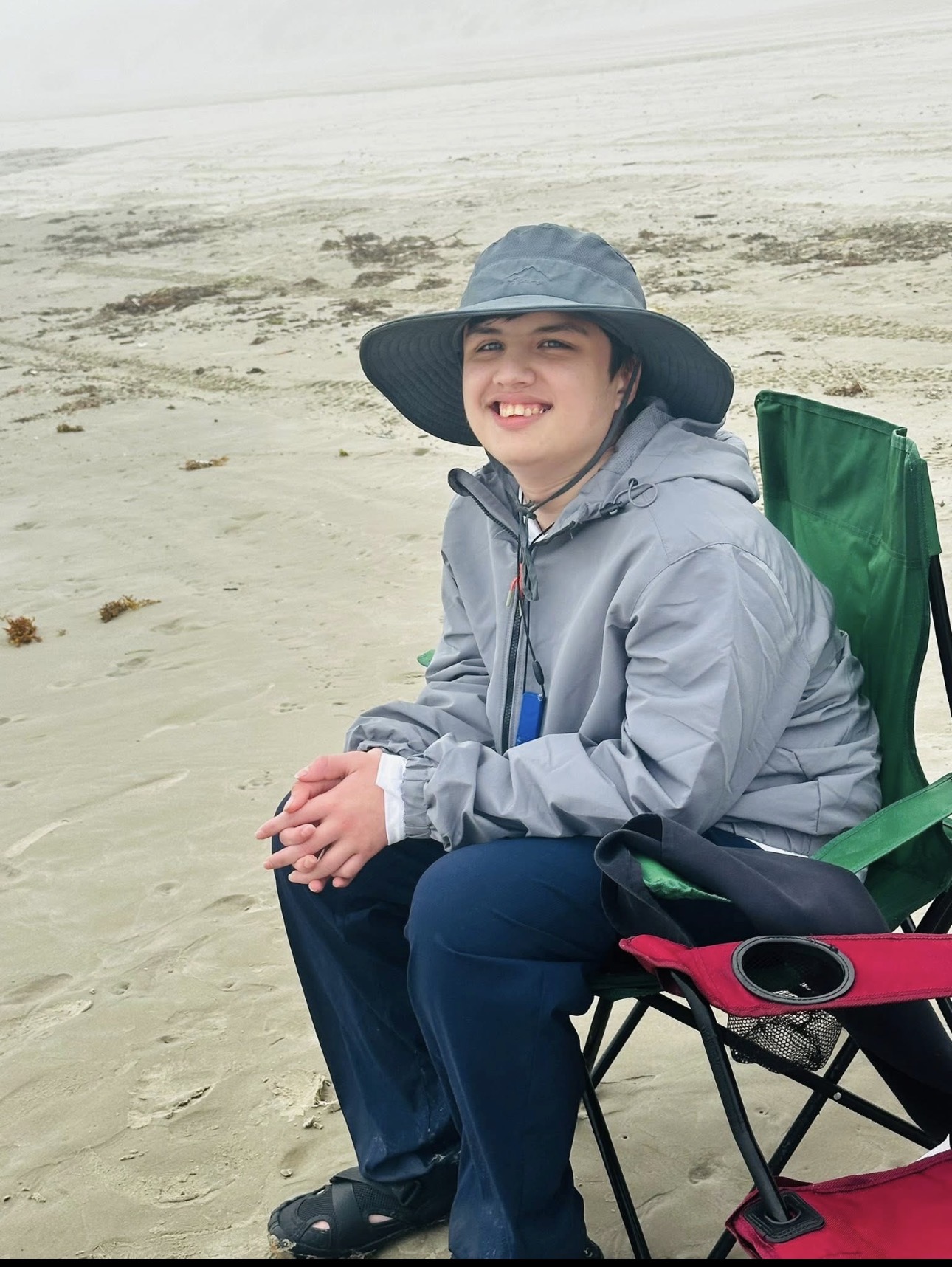 Noah smiling on the beach in a sun hat, sitting in a beach chair